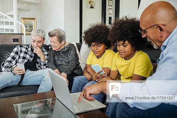 Zwillingsjungen mit Großvater  der einen Laptop benutzt  während ein junger Mann mit einem Smartphone bei der Urgroßmutter im Wohnzimmer sitzt