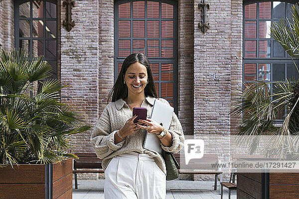 Lächelnde junge Frau mit Laptop und Smartphone auf dem Fußweg