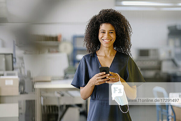 Smiling businesswoman with mobile phone and protective face mask standing in office