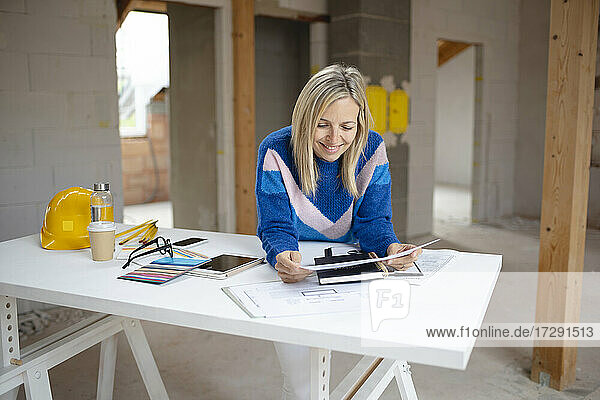Lächelnde Architektin  die einen Bauplan prüft  während sie sich in einer Wohnung auf einen Tisch stützt