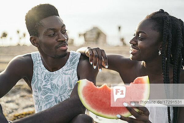 Smiling girlfriend with watermelon slice leaning on boyfriend shoulder at beach