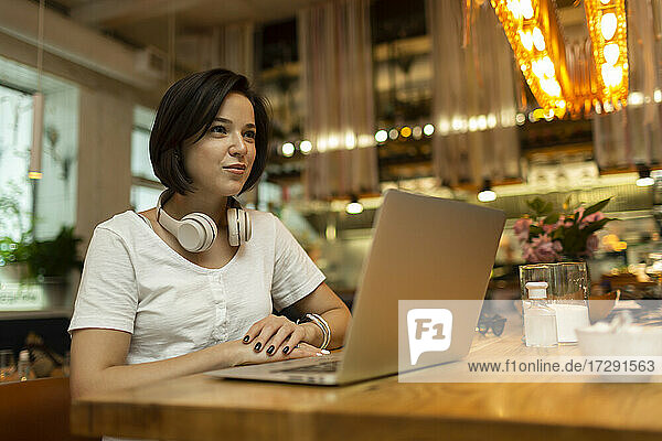 Lächelnde junge Freiberuflerin sitzt mit Laptop im Restaurant und schaut weg