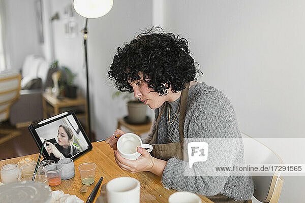 Künstler  der eine Tasse malt  während er durch ein Online-Tutorial auf einem digitalen Tablet am Arbeitsplatz lernt