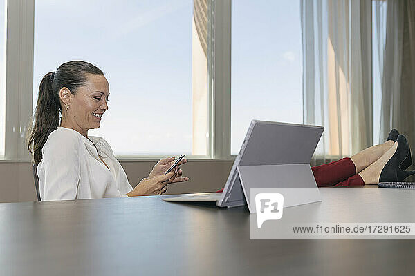Lächelnde Geschäftsfrau  die über ihr Smartphone Textnachrichten verschickt  während sie am Schreibtisch im Büro sitzt