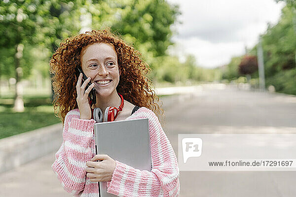 Happy redhead woman with curly talking on smart phone at park while looking away