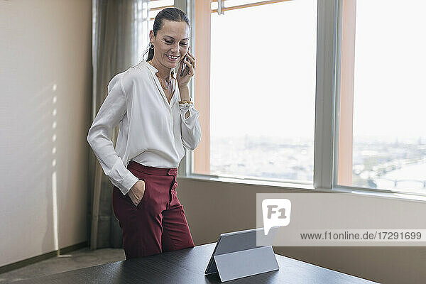 Lächelnde reife Geschäftsfrau  die auf einem Smartphone spricht  während sie am Schreibtisch im Büro steht