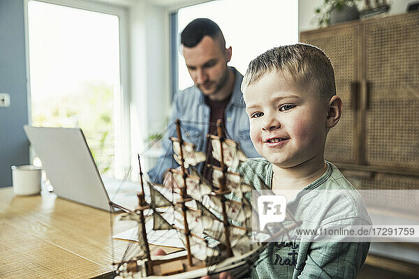 Cute boy playing with toy boat while father working on laptop at home