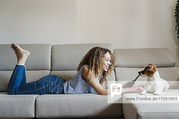 Lockenhaarige Frau streichelt Jack Russell Terrier  während sie auf dem Sofa im Wohnzimmer liegt