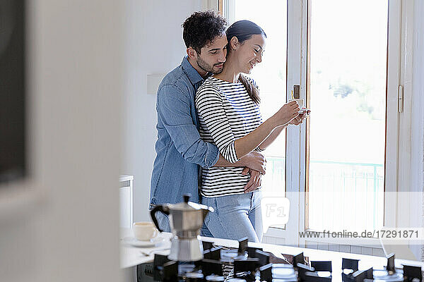 Boyfriend embracing smiling girlfriend from back in domestic kitchen