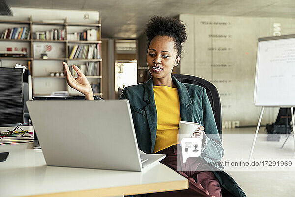 Geschäftsfrau mit Kaffeebecher gestikuliert während einer Videokonferenz am Laptop im Büro