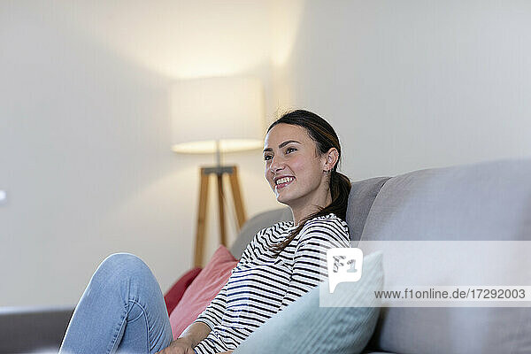 Smiling young woman day dreaming while sitting on sofa at home
