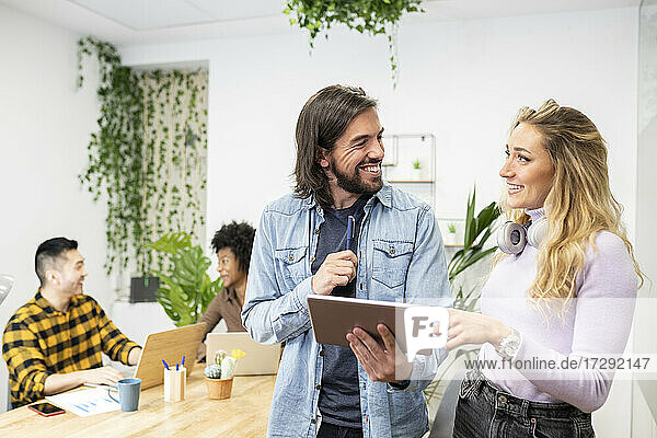 Cheerful male and female colleague looking at each other while discussing over digital tablet in office