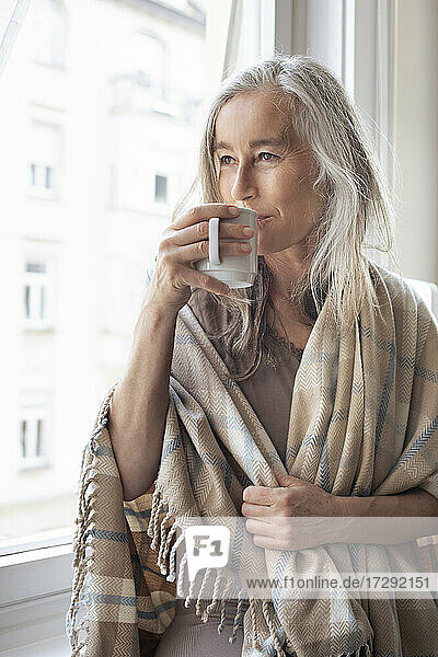 Reife Frau mit weißen Haaren schaut durch das Fenster und trinkt zu Hause Kaffee