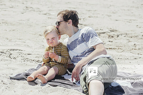 Father kissing son on head while sitting at beach during sunny day