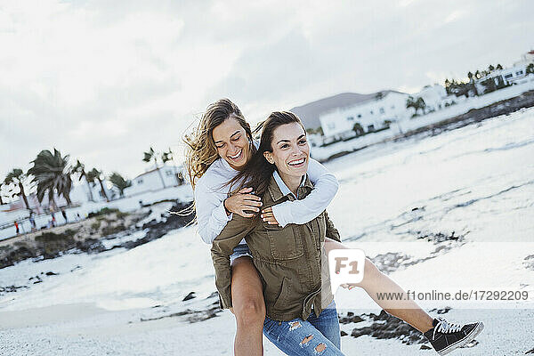 Playful woman piggybacking smiling friend on beach