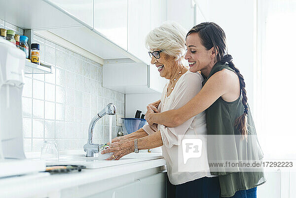 Lächelnde Frau im mittleren Erwachsenenalter  die ihre Großmutter umarmt  die zu Hause in der Küche Geschirr wäscht