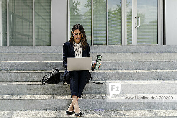 Geschäftsfrau hält Reisetasse und benutzt Laptop  während sie auf einer Treppe sitzt