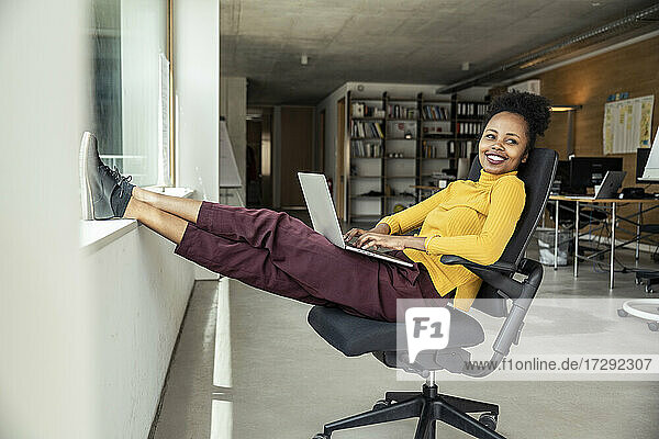 Lächelnde weibliche Fachkraft mit Laptop schaut weg  während sie auf einem Stuhl im Büro sitzt