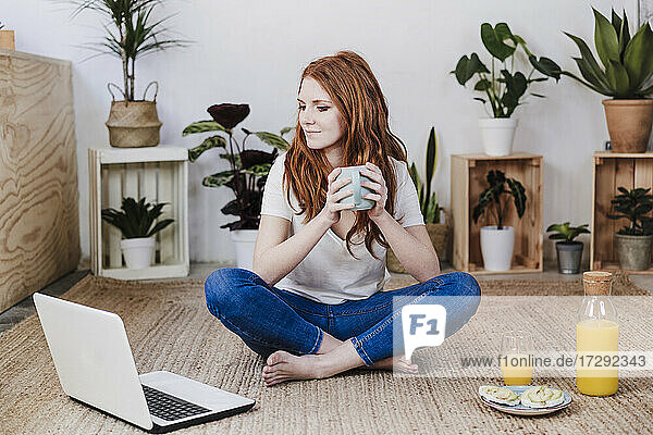 Frau mit gekreuzten Beinen  die auf einen Laptop schaut  während sie einen Kaffee auf dem Boden zu Hause trinkt