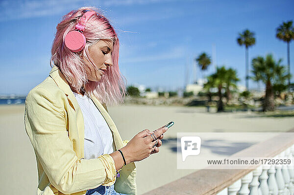 Frau mit rosafarbenem Haar  die an einem sonnigen Tag an der Promenade ein Mobiltelefon benutzt