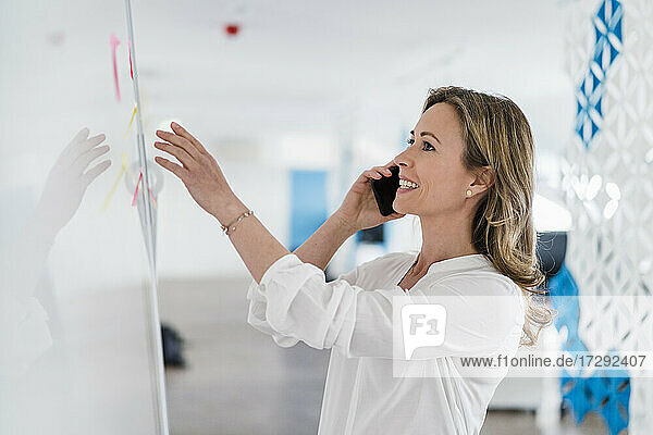 Lächelnde weibliche Fachkraft im Gespräch auf dem Smartphone vor dem Whiteboard im Büro