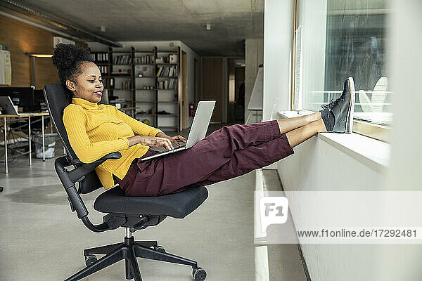 Weibliche Fachkraft  die einen Laptop benutzt  während sie mit hochgelegten Füßen auf einem Stuhl im Büro sitzt