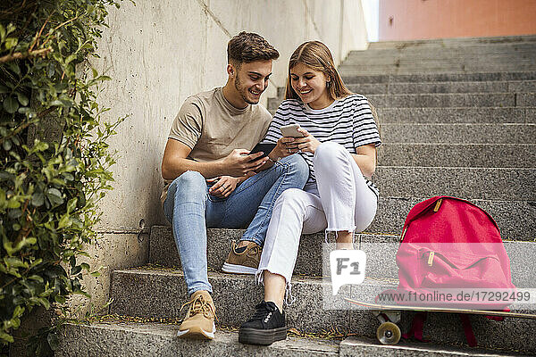 Lächelndes Paar  das auf einer Treppe sitzend ein Mobiltelefon benutzt