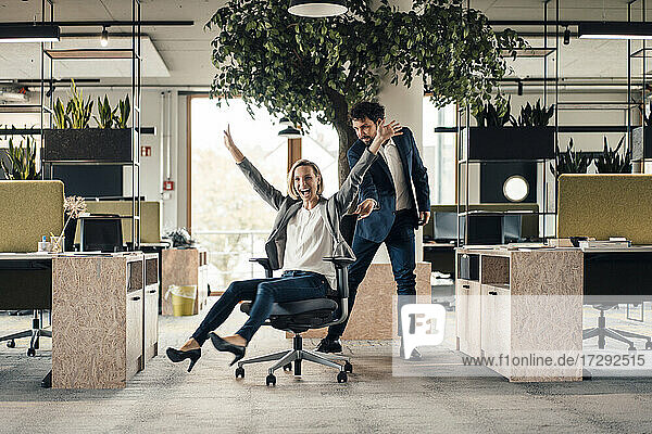 Fröhliche Geschäftsfrau auf einem Stuhl sitzend mit einem im Hintergrund stehenden Kollegen im Büro