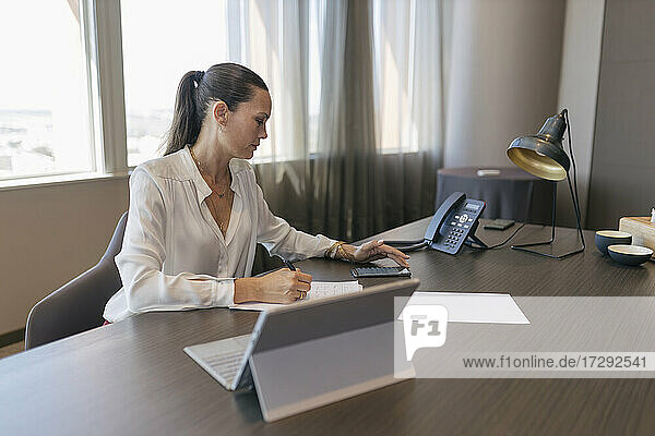 Weibliche Fachkraft  die ein Smartphone benutzt  während sie an einem Dokument am Schreibtisch im Büro schreibt