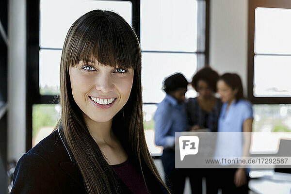Smiling beautiful businesswoman with bangs standing while male and female colleagues discussing in office