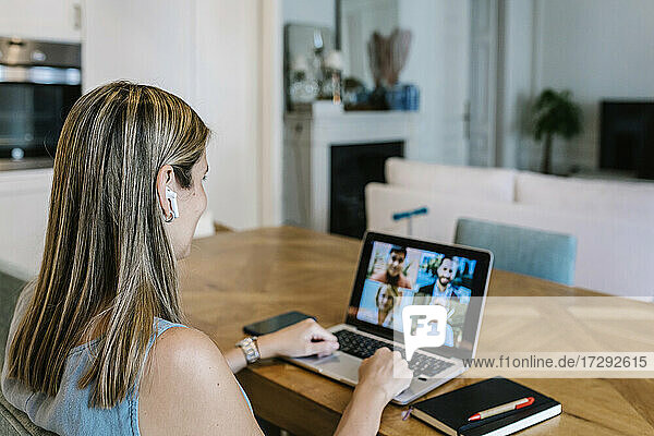 Female freelance worker wearing wireless earphones talking on video conference through laptop at home
