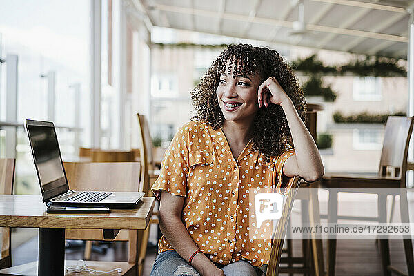 Lächelnde Geschäftsfrau mit Kopf in der Hand träumt  während sie in einem Café am Laptop sitzt