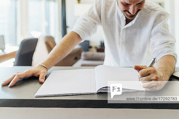 Smiling young man writing on book at kitchen island