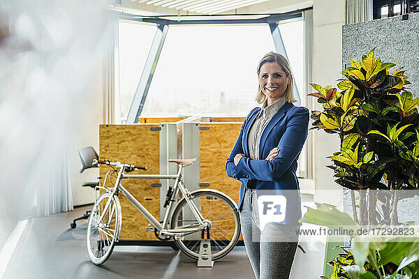 Lächelnde Unternehmerin steht mit verschränkten Armen neben einer Topfpflanze in einem kreativen Büro