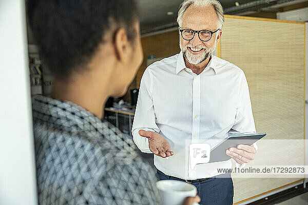 Lächelnder Geschäftsmann mit digitalem Tablet gestikuliert  während er eine Kollegin im Büro ansieht