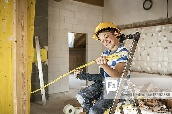 Lächelnder Junge schaut auf ein Maßband  während er auf einer Leiter auf einer Baustelle sitzt