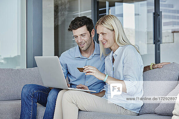 Smiling couple using laptop while doing online shopping sitting in living room at home
