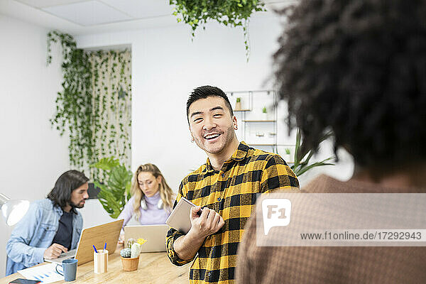 Lächelnder junger Geschäftsmann mit digitalem Tablet  der eine Mitarbeiterin beim Diskutieren im Büro ansieht
