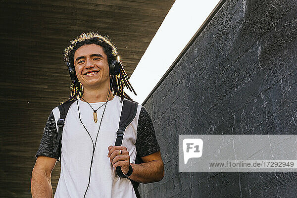 Lächelnder Mann hört Musik über Kopfhörer  während er einen Rucksack an der Wand trägt
