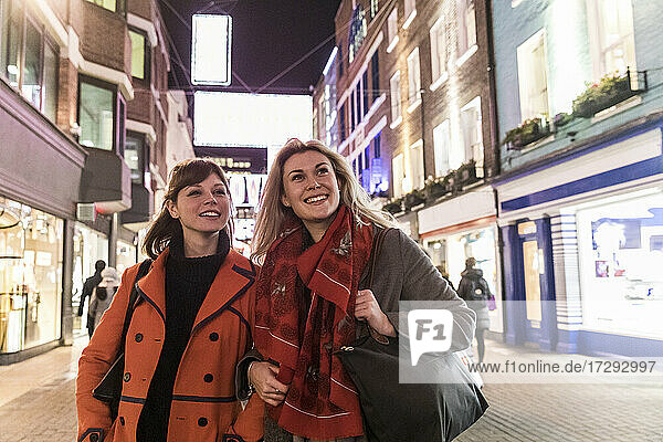Lächelnde Freundinnen in Wintermänteln schauen weg  während sie zusammen in der nächtlichen Stadt spazieren gehen