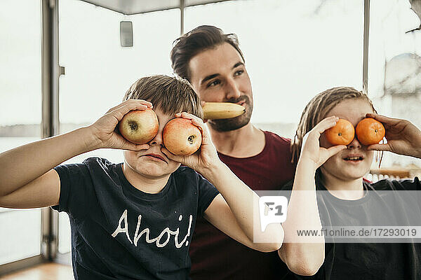 Verspielte Brüder bedecken ihre Augen mit Früchten  während sie mit ihrem Vater in der Küche sitzen