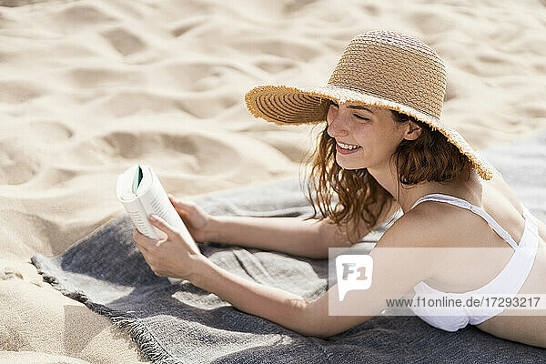 Lächelnde Frau mit Hut  die wegschaut  während sie in den Ferien auf einem Handtuch am Strand liegt