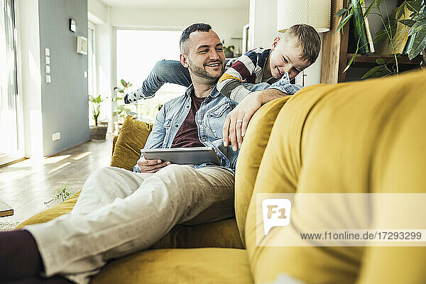 Lächelnder Mann  der mit einem Tablet sitzt und seinen Sohn beim Spielen im Wohnzimmer beobachtet