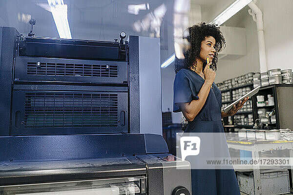 Nachdenkliche Geschäftsfrau mit digitalem Tablet  die wegschaut  während sie an einer Maschine in der Industrie steht