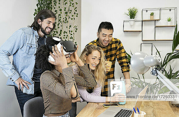 Cheerful businesswoman wearing virtual reality simulator by male and female coworkers doing video call through laptop in office