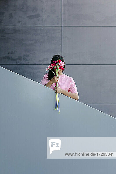 Junge Frau versteckt ihr Gesicht mit Gerbera-Gänseblümchen vor einem Geländer