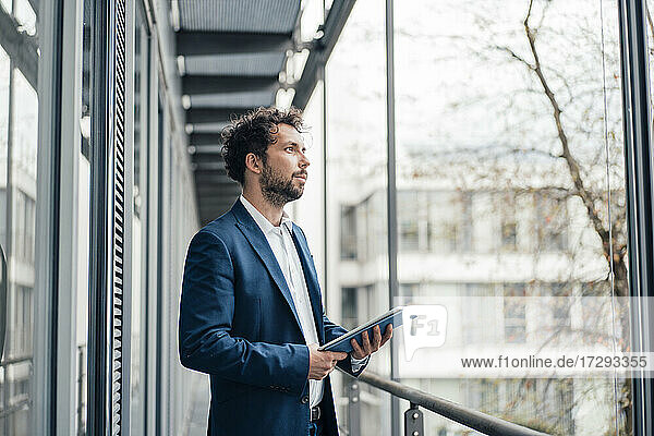 Nachdenklicher Geschäftsmann mit digitalem Tablet  der am Fenster steht und wegschaut