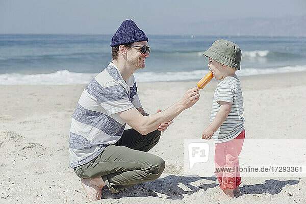 Smiling man feeding ice cream to son at beach during sunny day