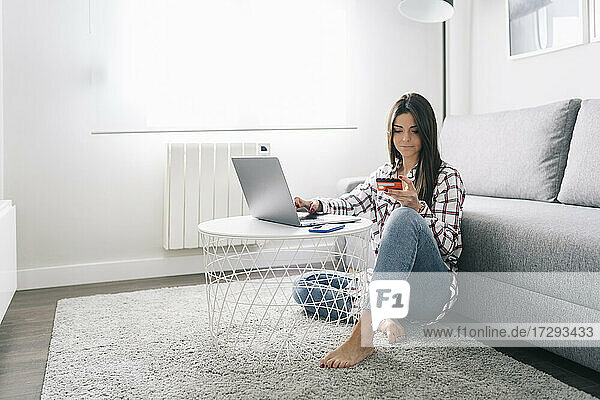Junge Frau mit Kreditkarte beim Online-Shopping mit Laptop im Wohnzimmer