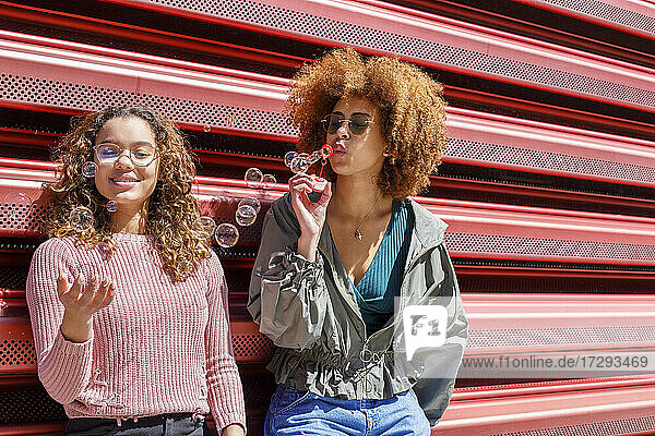 Junge Freundinnen spielen mit Seifenblasen vor einer Wellblechwand an einem sonnigen Tag